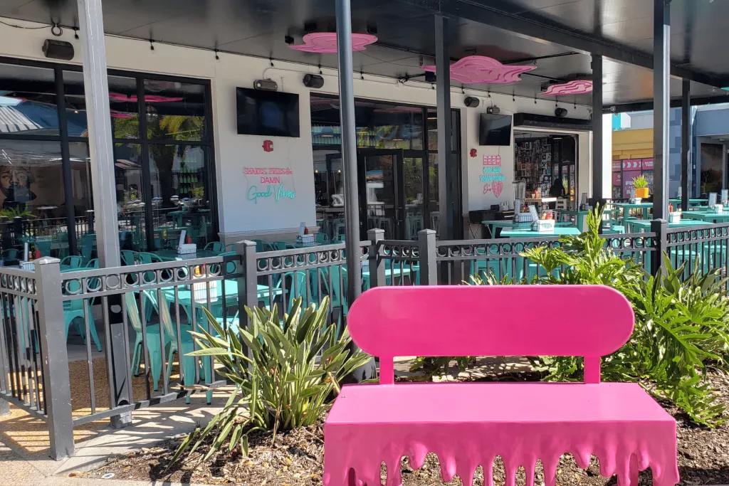 A covered outdoor patio at a restaurant with blue tables and chairs and a bright pink bench in the foreground