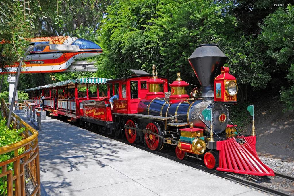 A classic steam train with red accents waiting in a station with a monorail going past in the background