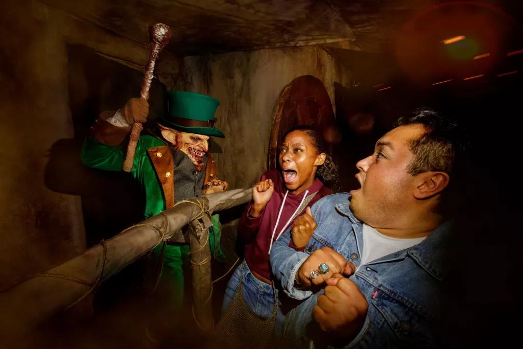 A scare actor dressed as a leprechaun jumping out at a man and a woman who are both screaming  
