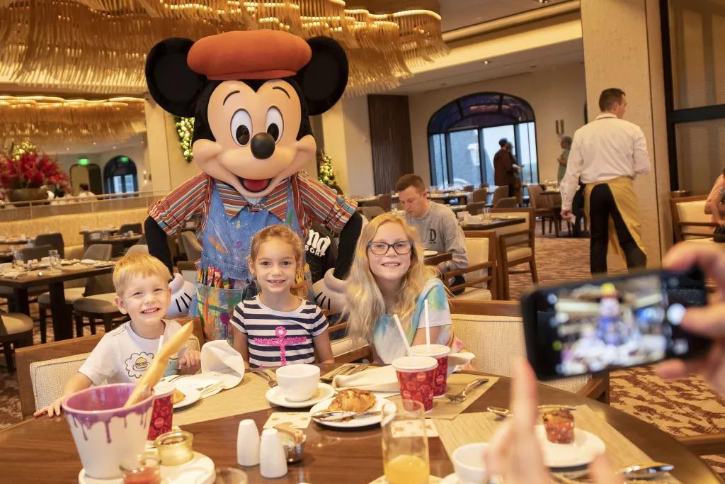 Three children sat at a table with Mickey Mouse posing behind them