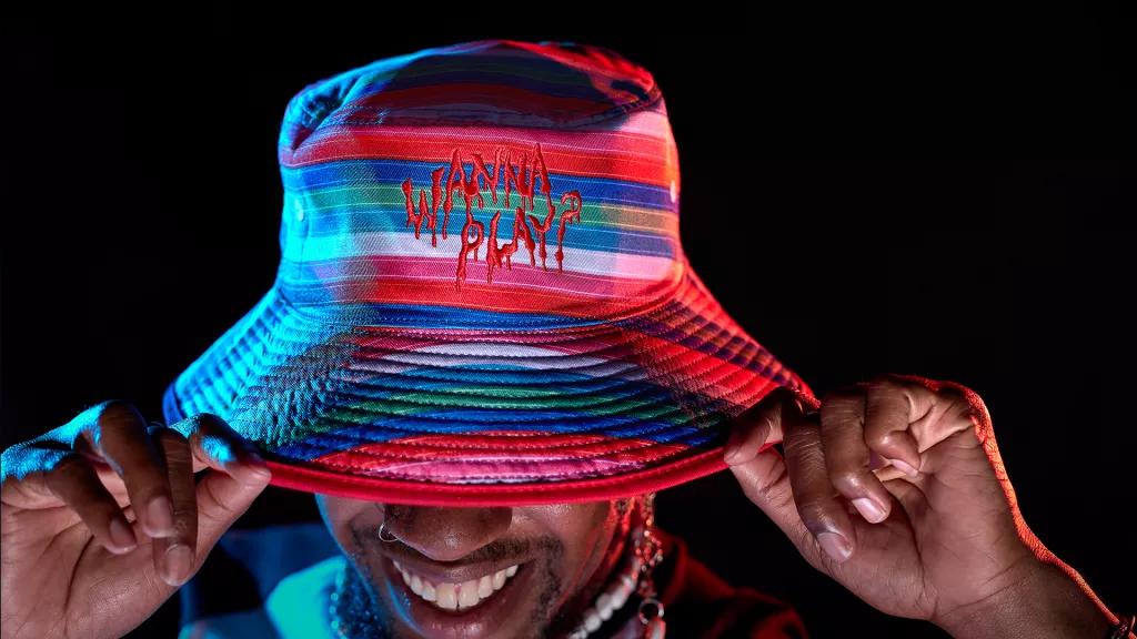 A man looking down holding the brim of a striped bucket hat that read's 'Wanna Play?'