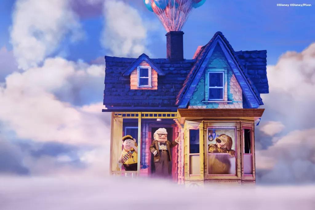 Carl, Russel, and Dug standing in front of a multicoloured house on a stage covered with fog