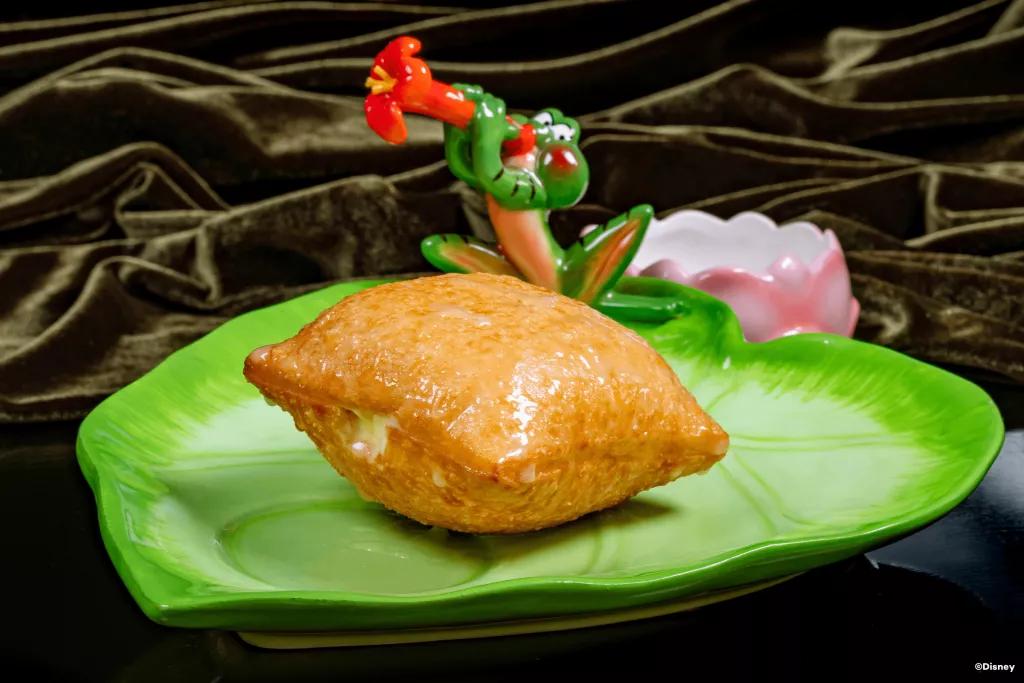 A filled and glazed beignet on a plate shaped like a lily pad with a frog playing a flower like a trumpet