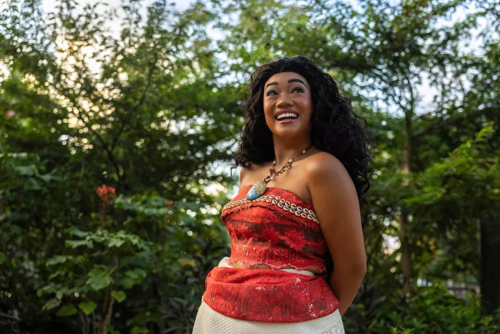 Moana standing with her hands behind her back in front of a group of trees 