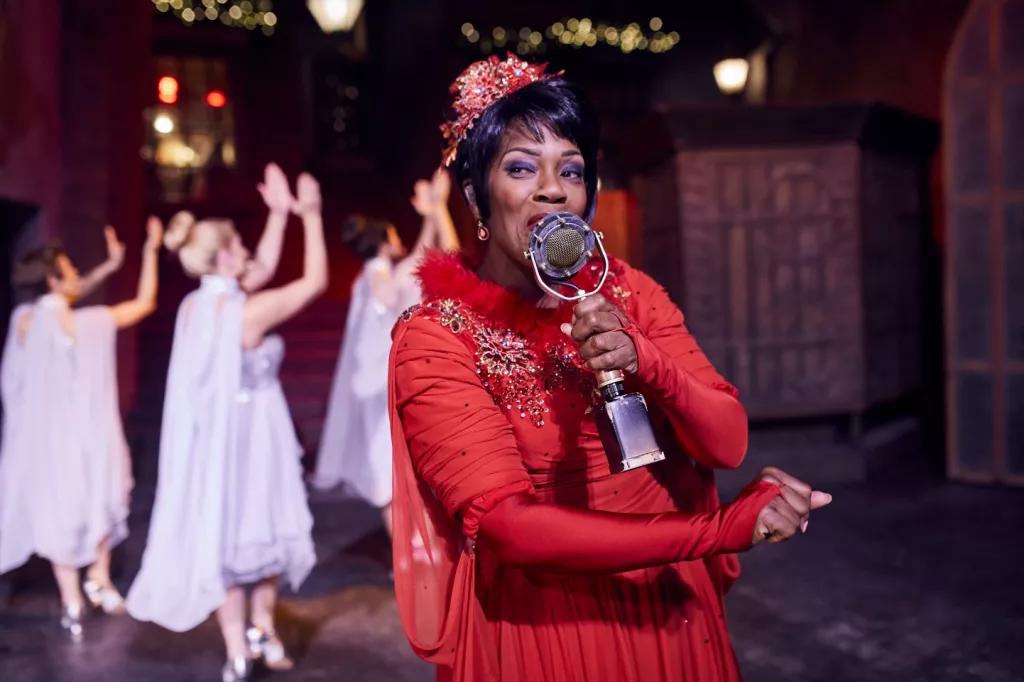 A woman in a red dress singing into an old fashioned microphone. Three women in white dresses are dancing behind her