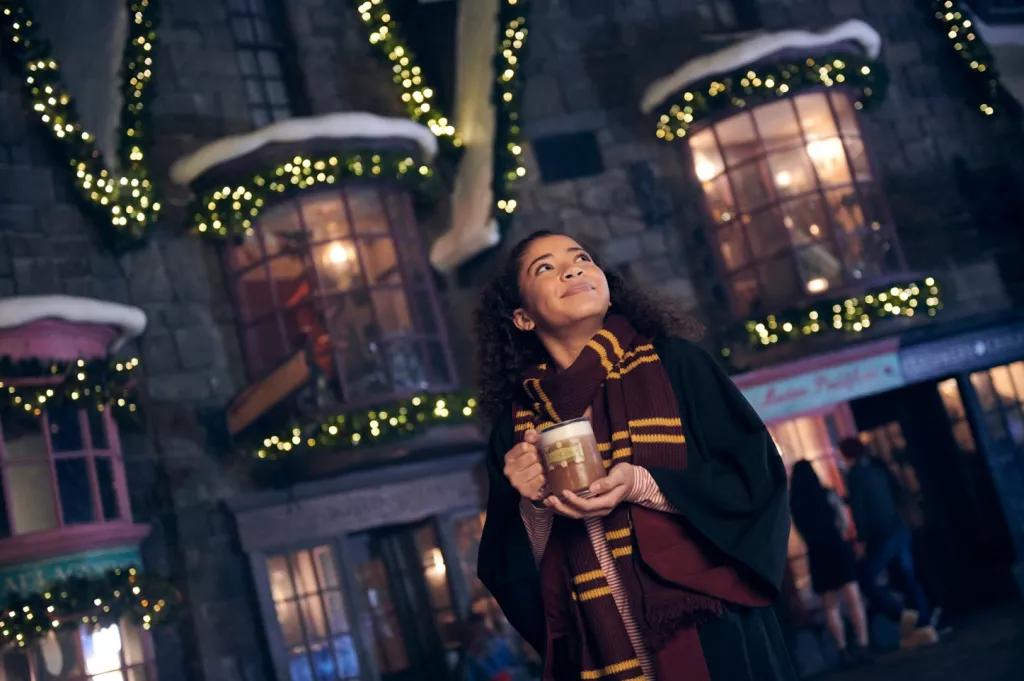 A woman wearing a black Hogwarts cloak and Gryffindor scarf looking up while holding a mug of warm Butterbeer