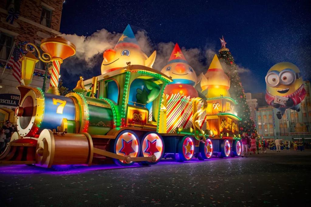 A parade float shaped like a giant toy train lit up with lights. A large balloon shaped like a Minion follows