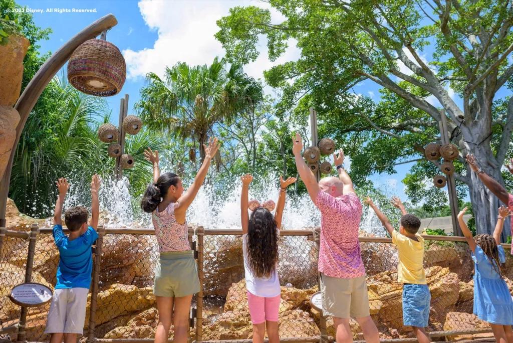 A family standing in a line facing away from the camera holding their arms up to control the water fountains in front of them 
