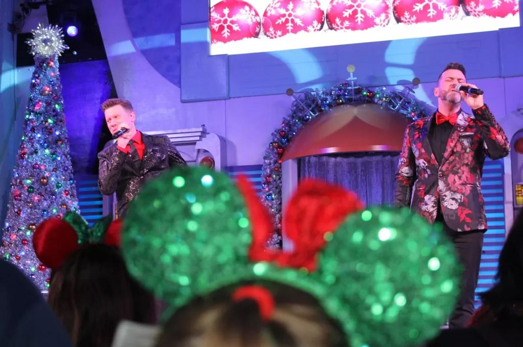 Two men singing on a stage wearing red and black suits. A pair of sparkly green and red Minnie ears is in the foreground