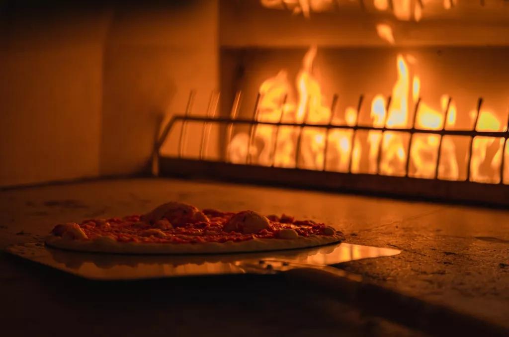 A thin crust pizza being put into a pizza oven with flames in the background