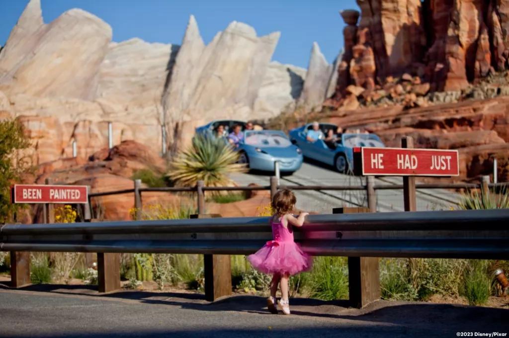 A young girl in a pink tutu watching two ride vehicles go past designed to look like cars from the Pixar movie Cars
