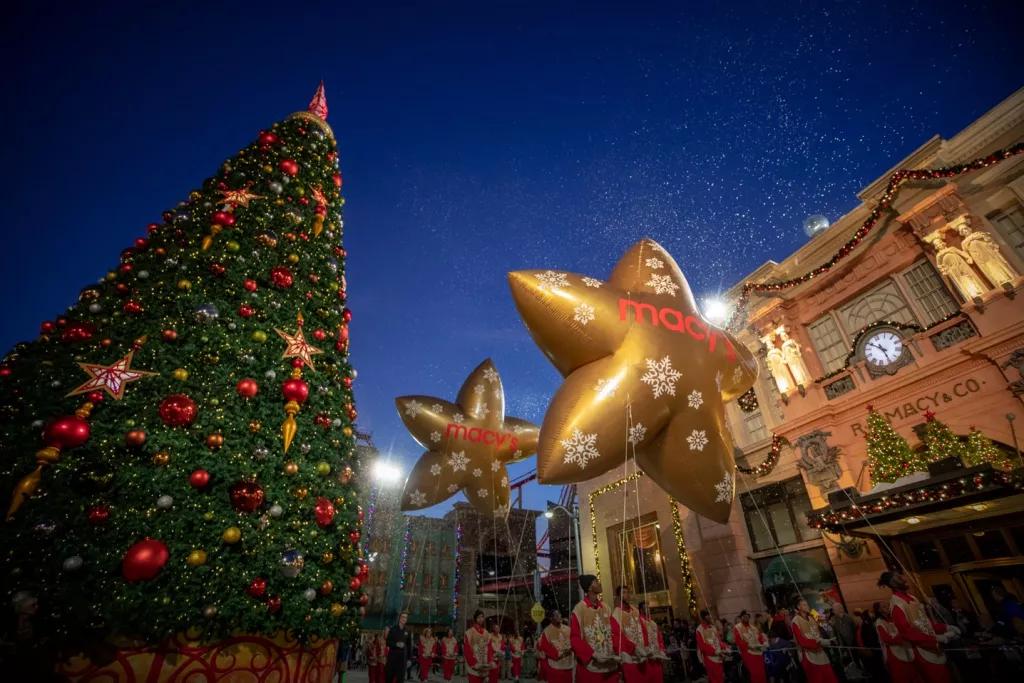 Large gold star-shaped balloons being carried past a tall Christmas tree