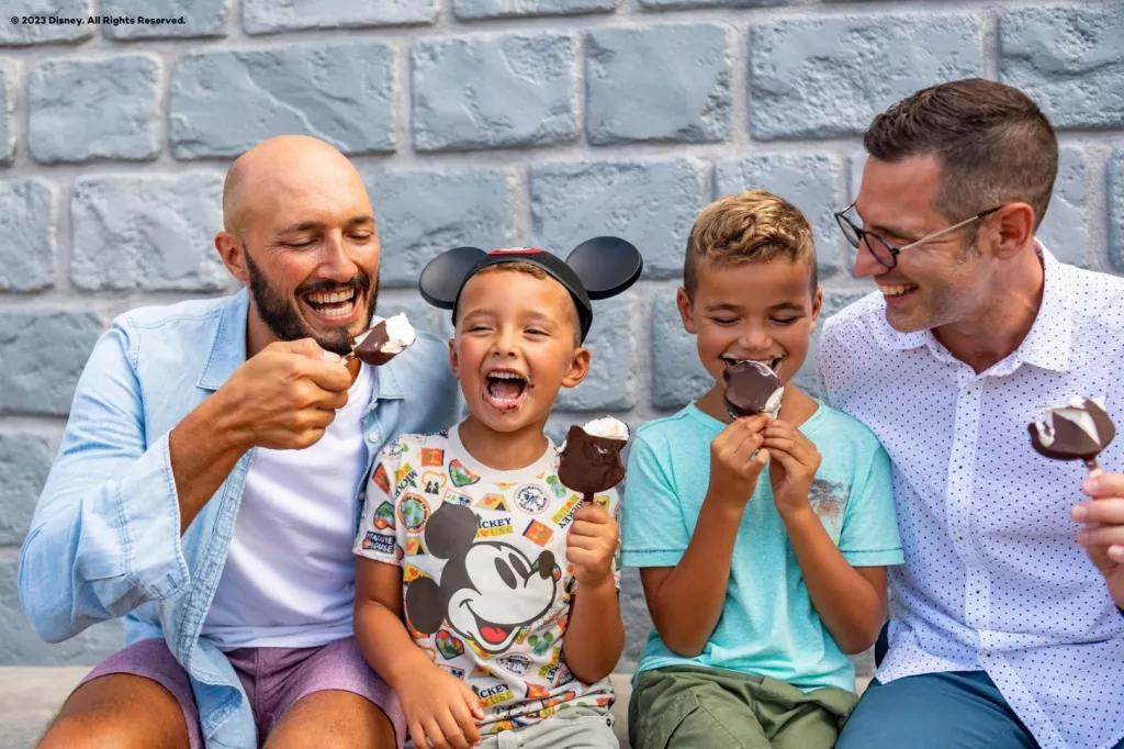 Two men and two young boys sat down eating chocolate covered ice cream