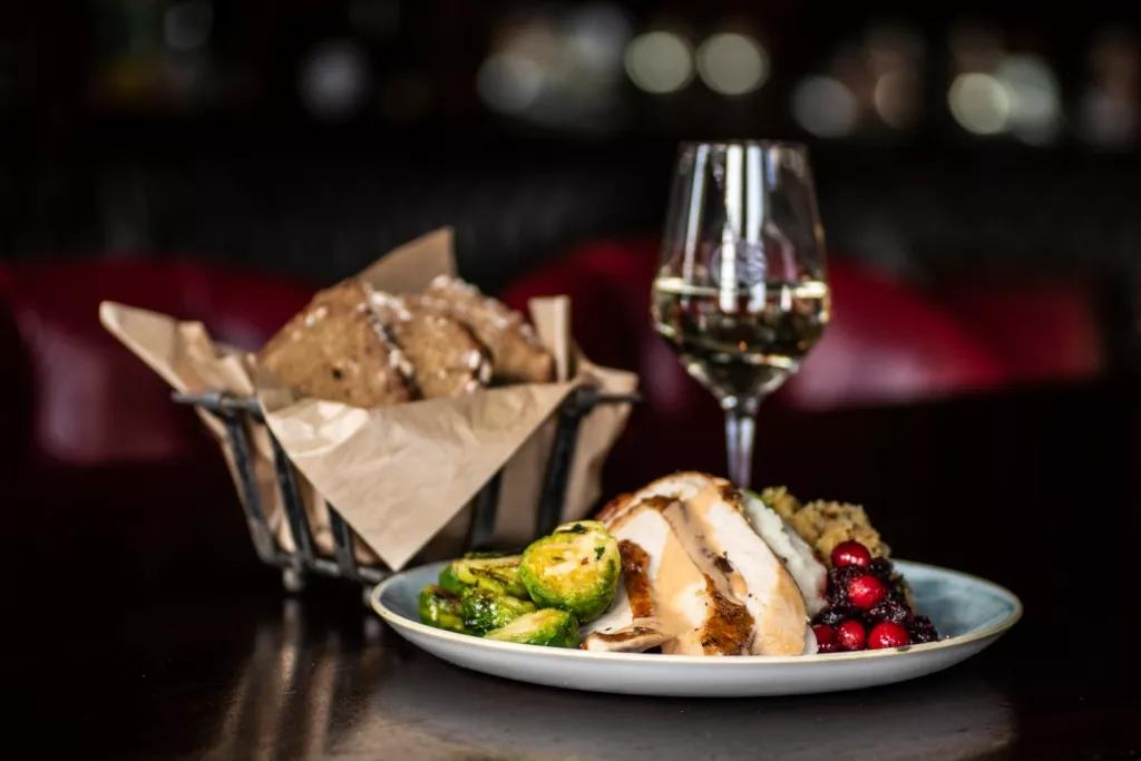 A plate of turkey, brussel sprouts, stuffing and cranberry sauce in front of a basket of bread and a glass of wine