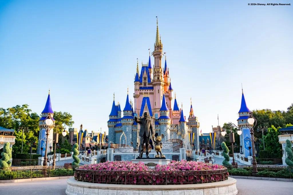 A large pink and blue castle in front of a clear blue sky. A statue of Walt Disney and Mickey Mouse is in the foreground