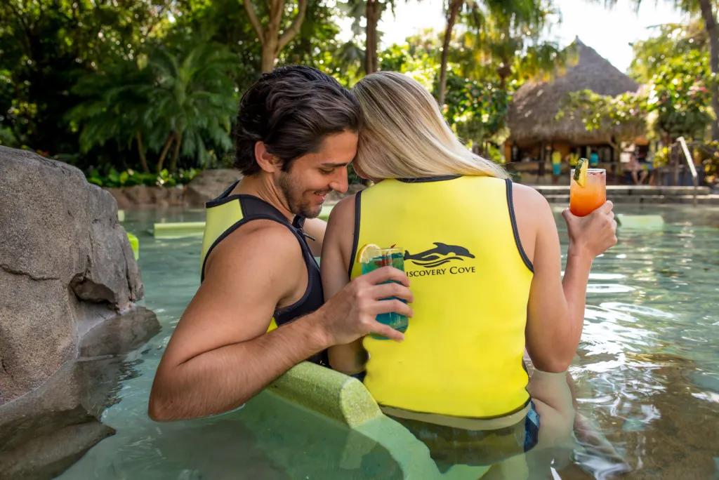 A couple sitting in shallow water holding cocktails and leaning in to each other