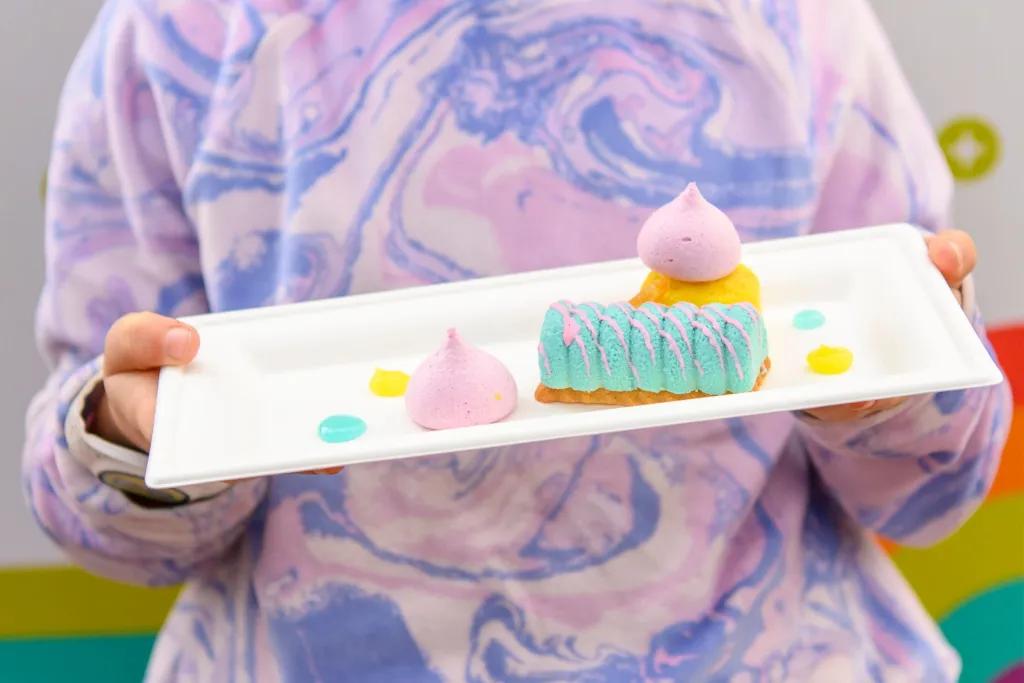 Someone wearing a purple tie-dye jumper holding a plate of colourful desserts