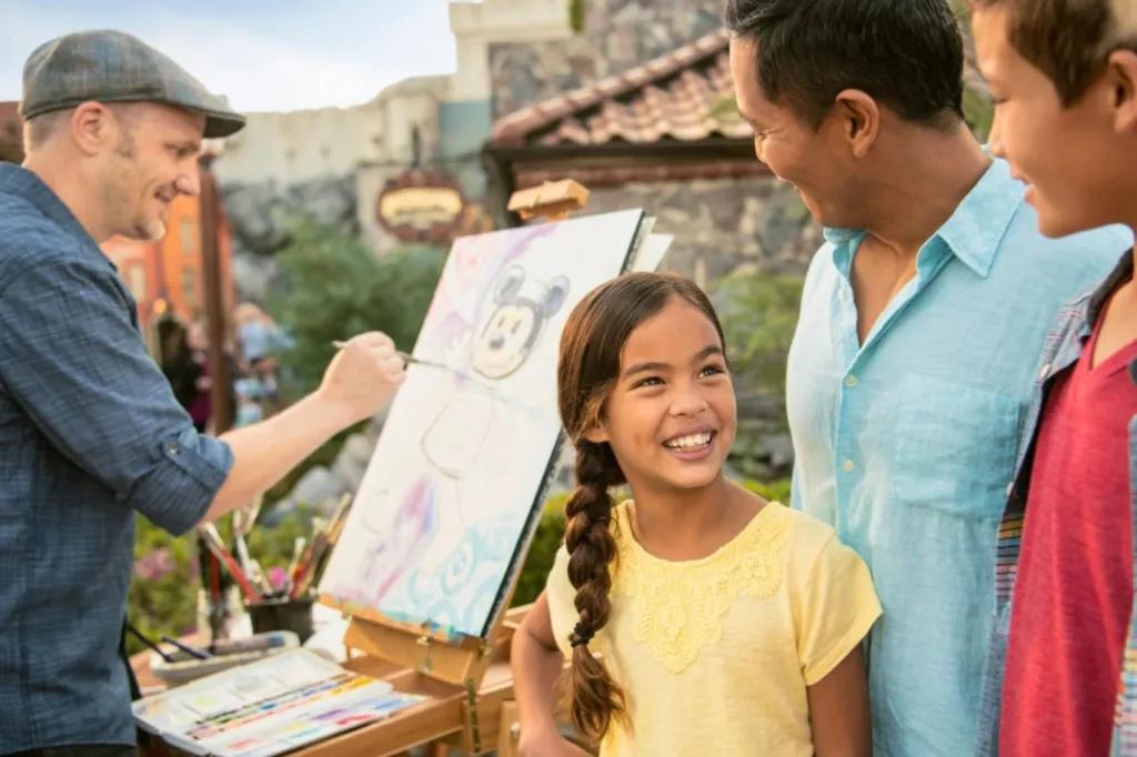 A family watching a man paint a stylised picture Mickey Mouse on a canvas sat on an easel