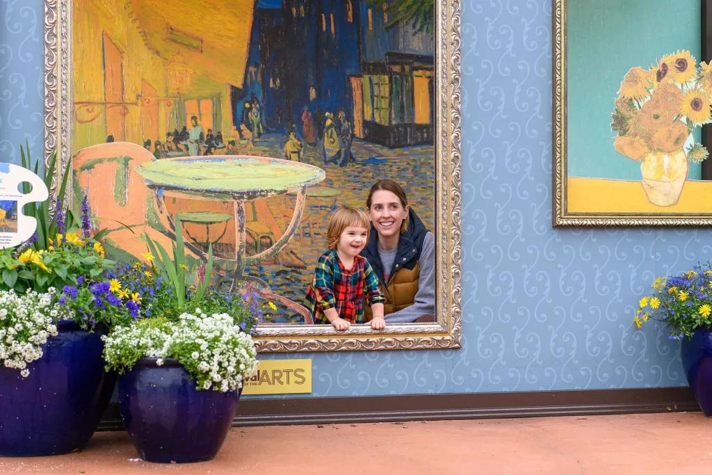 A woman and young boy posing inside a large picture frame in front of a background designed to look like they are in a painting