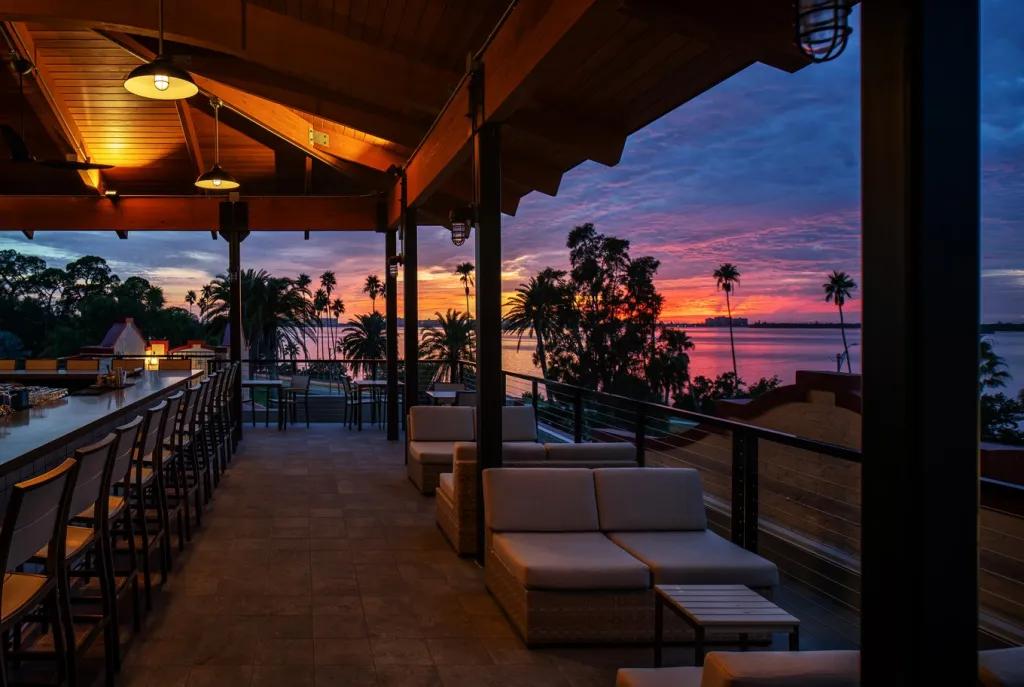 An outdoor dining terrace with low tables and bar seating. In the background is a blue and pink sunset over a beach