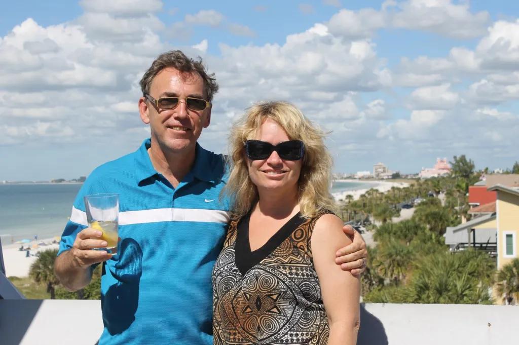 A man and woman posing on a balcony in front of a shoreline lined with palm trees