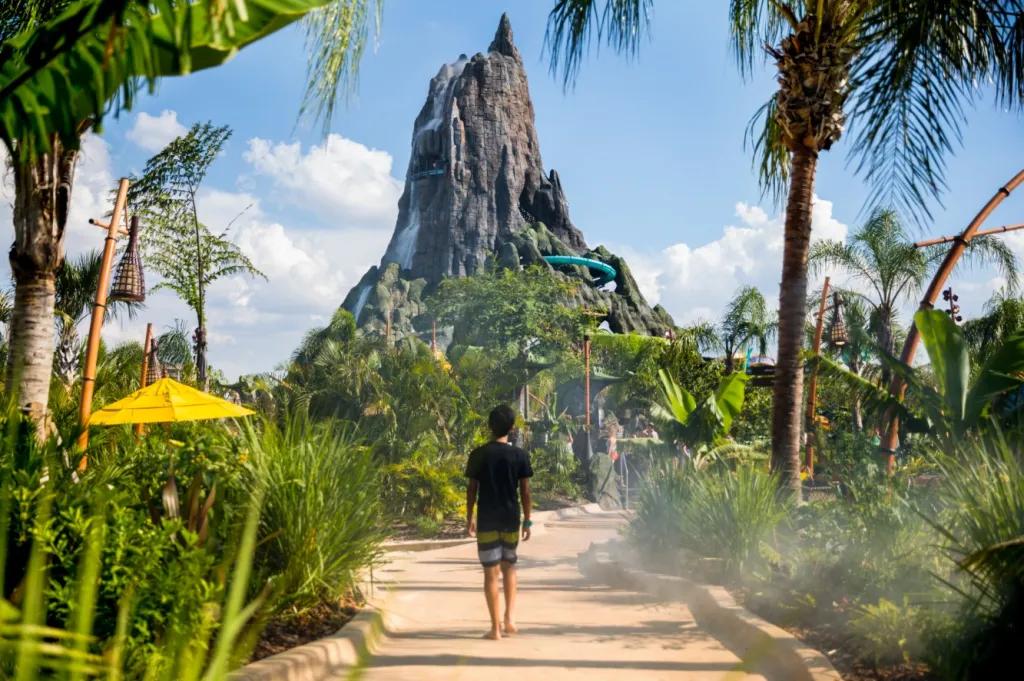 A boy wearing a tshirt and shorts walking along a path surrounded by greenery towards a volcano spouting water