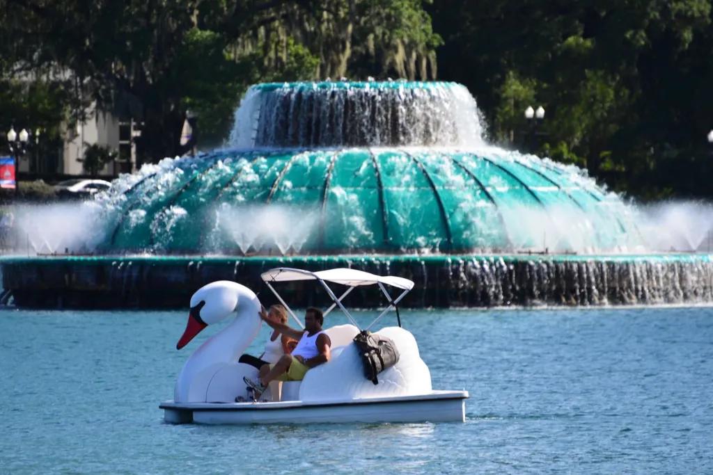 Two people in a swan shaped peddle boat on a lake in front of a large circular fountain