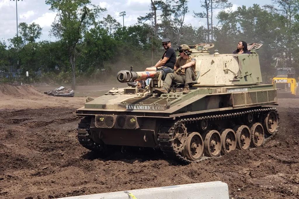 An army tank being driven across soil with two people sitting at the front and one person at the back