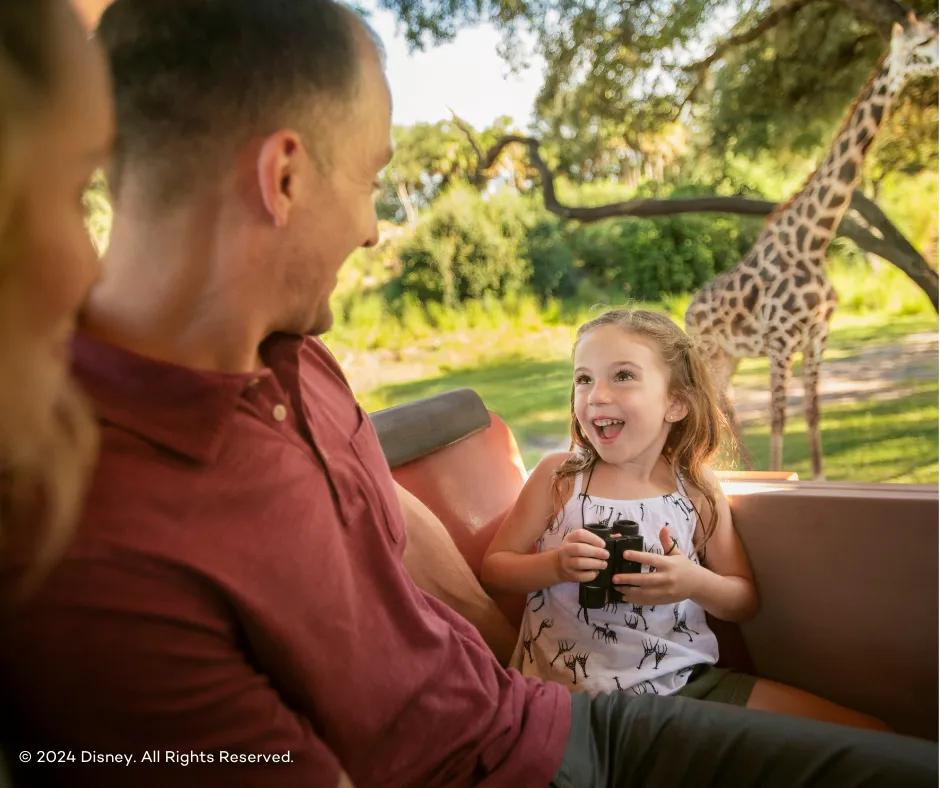 Small girl holding binoculars in truck facing her parents with giraffe stood behind her eating leaves in a tree and her dad sat to the left of her smiling down at her.