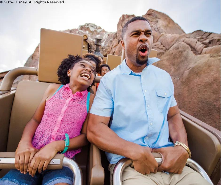 A young girl and an older man sat together on the ride with the girl in a bright pink top leaning over smiling towards the man who is grabbing the lap restraints and screaming