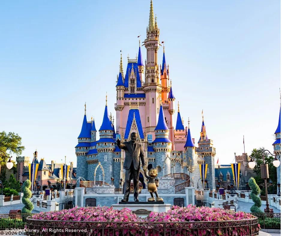Statue of Walt Disney holding hands with Mickey Mouse in front of large pink and blue castle