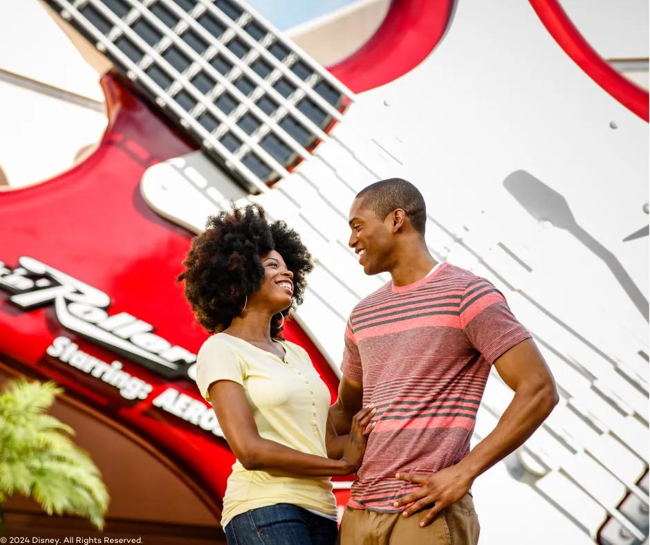 Couple looking at one another smiling, stood in front of a giant red guitar sign at the front of the Rock 'n' Roller Coaster ride with the ride title written on the bottom left of the guitar.