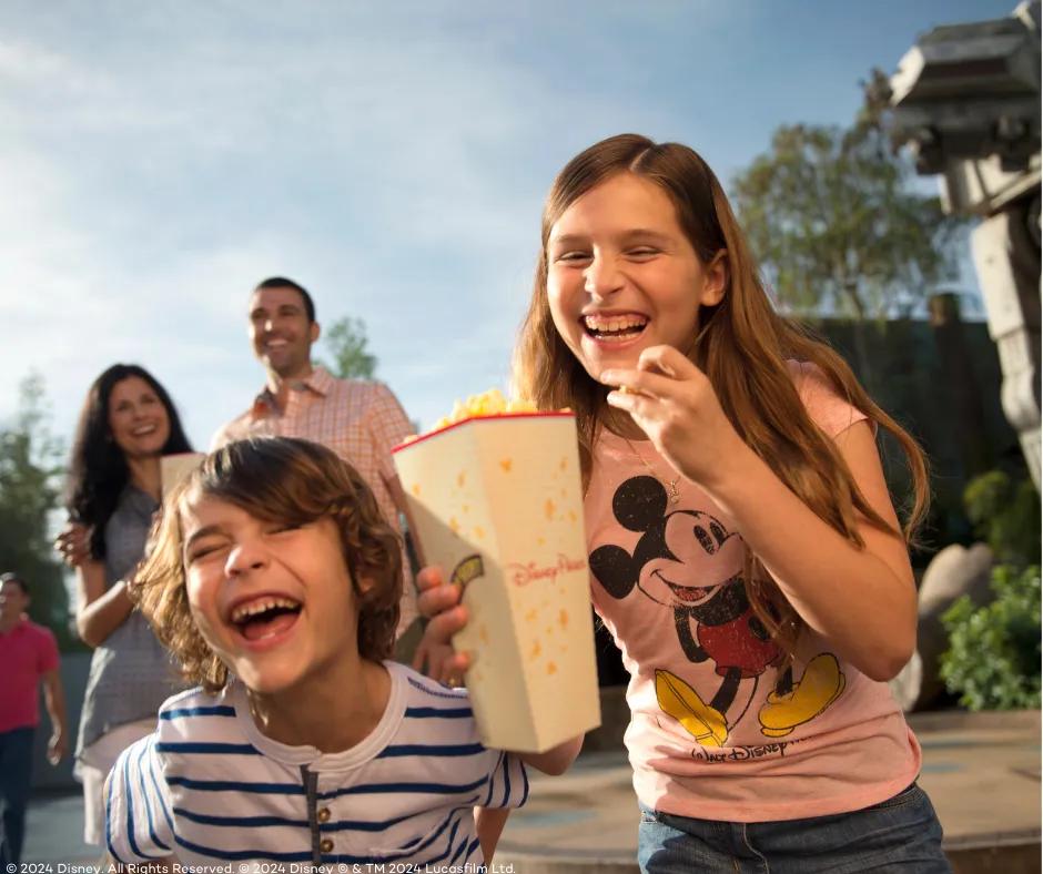 Girl wearing pink Mickey Mouse top holding popcorn bucket in one hand and a piece of popcorn in the other, laughing with a young boy who is next to her. In the background is a couple smiling and a large star wars robot.