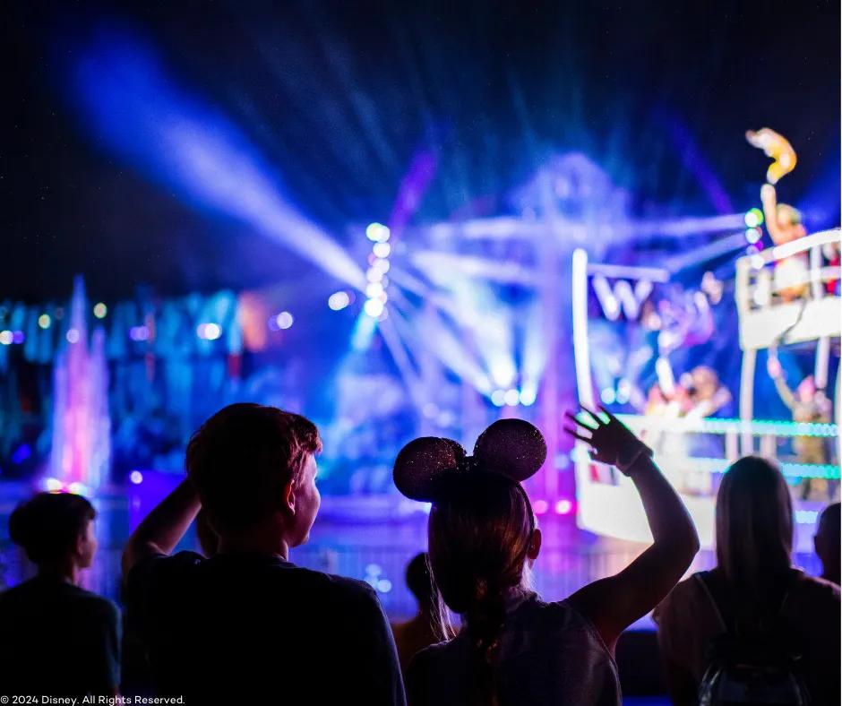 Behind two children watching Fantasmic! One girl wearing mickey ears and one boy with one of each of their hands in the air. In the blurry background there are multicoloured lasers shining, purple and blue lighting, fountains and the front of a white ferry boat holding Disney characters.
