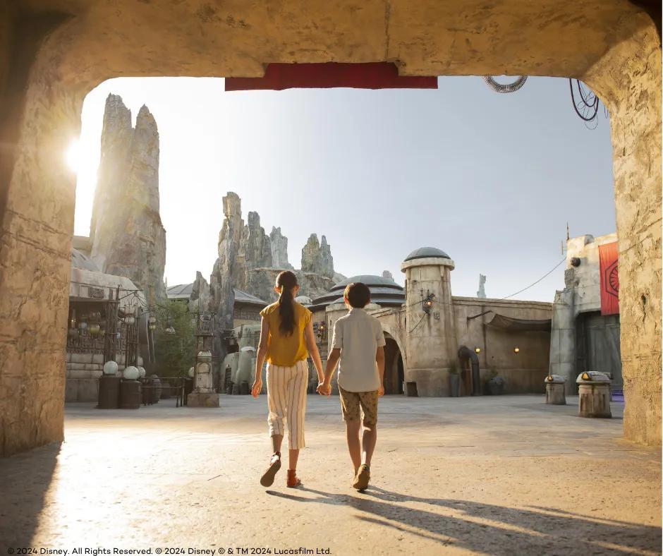Young girl and young boy holding hands walking with backs towards the camera, into area themed with grey large rocks and beige dirty buildings with circular roofs. There are wires hanging between the buildings.