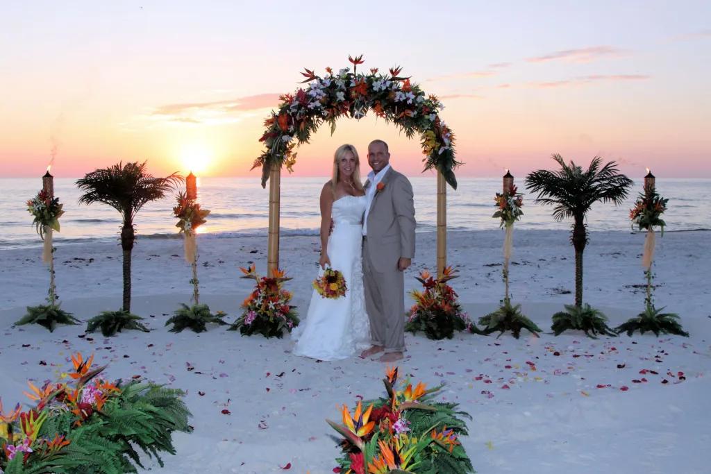 A bride and groom standing on a beach at sunset in front of a floral wedding arch