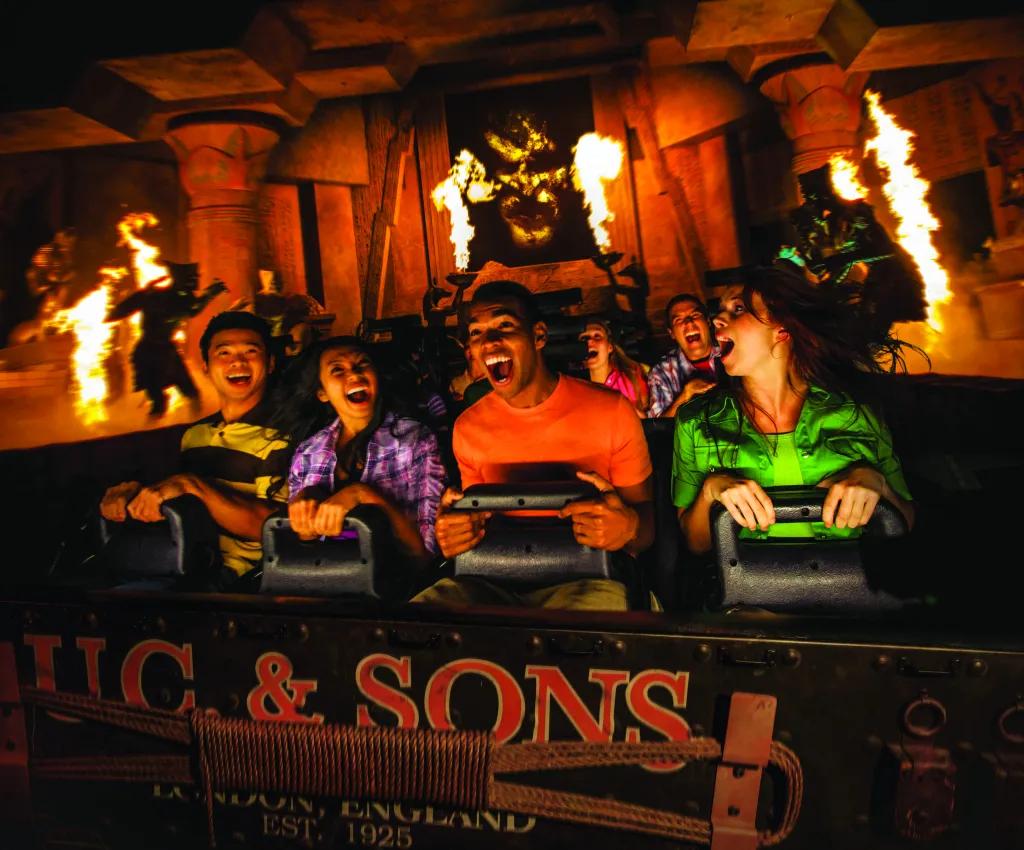 Four people laughing and smiling in shock on the mummy ride with fire pyrotechnics in the background and statues of mummies. A large scary face is projected in the back. 