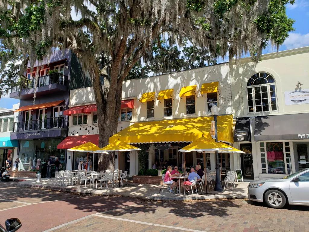 Row of cream coloured buildings with multicoloured shops and restaurants alongside. The central restaurant (Briarpatch) has bright yellow umbrellas outside with white chairs and tables.