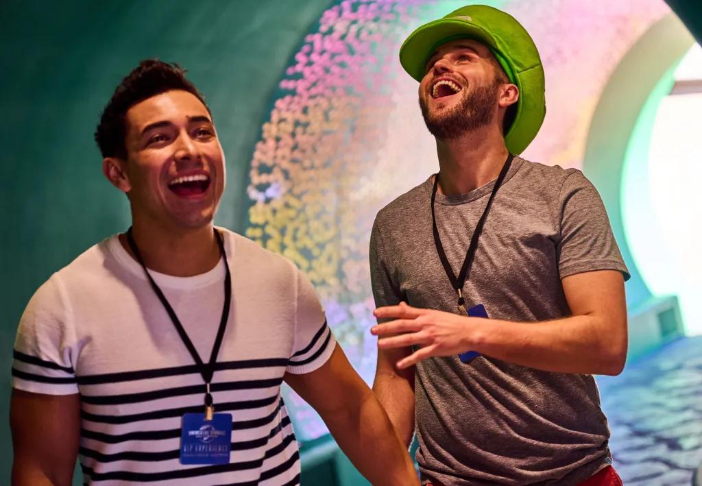 Two men walking through green tunnel with multicoloured lights on the ceiling. Both laughing and wearing lanyards, with one wearing a large green Luigi hat.