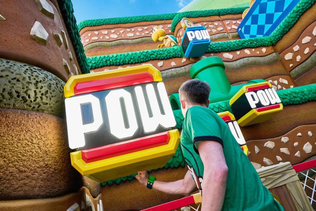 Man wearing green shirt and green power up band punching underneath a large box which is floating that says 'POW' in white writing. In the background is a brown hill with green grass style decorations, green pipes and small turtle shaped characters.