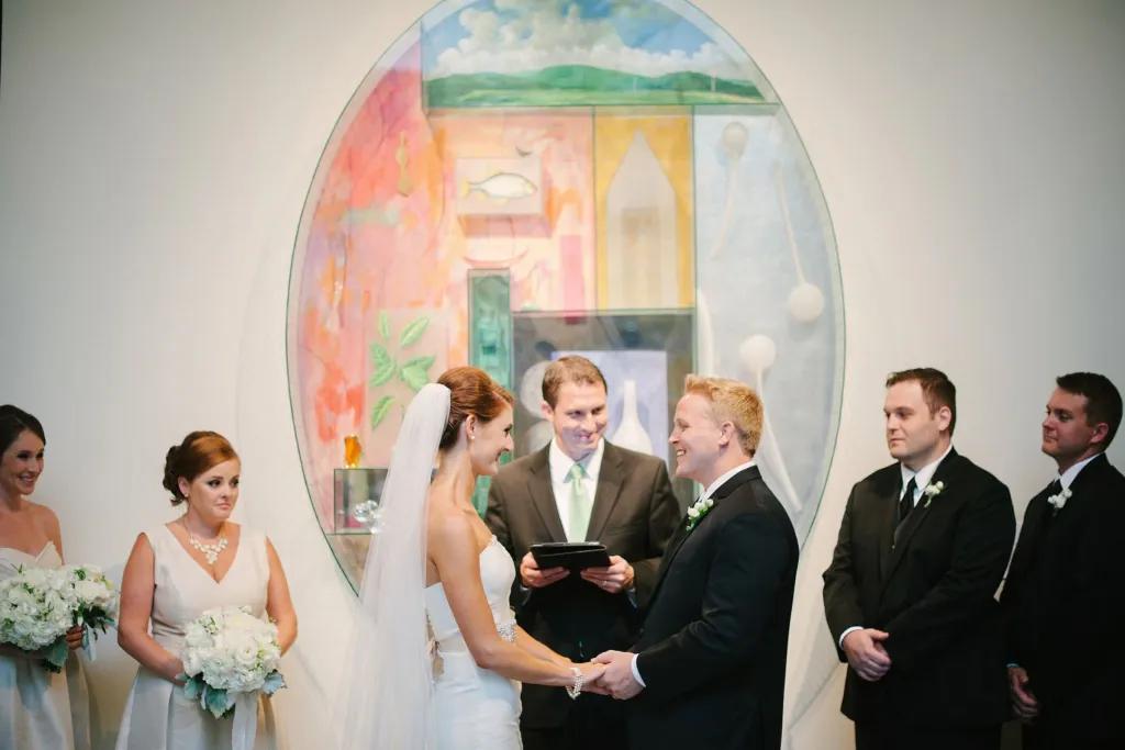 A bride and groom next to their bridesmaids and groomsmen getting married in front of a large oval-shaped piece of art