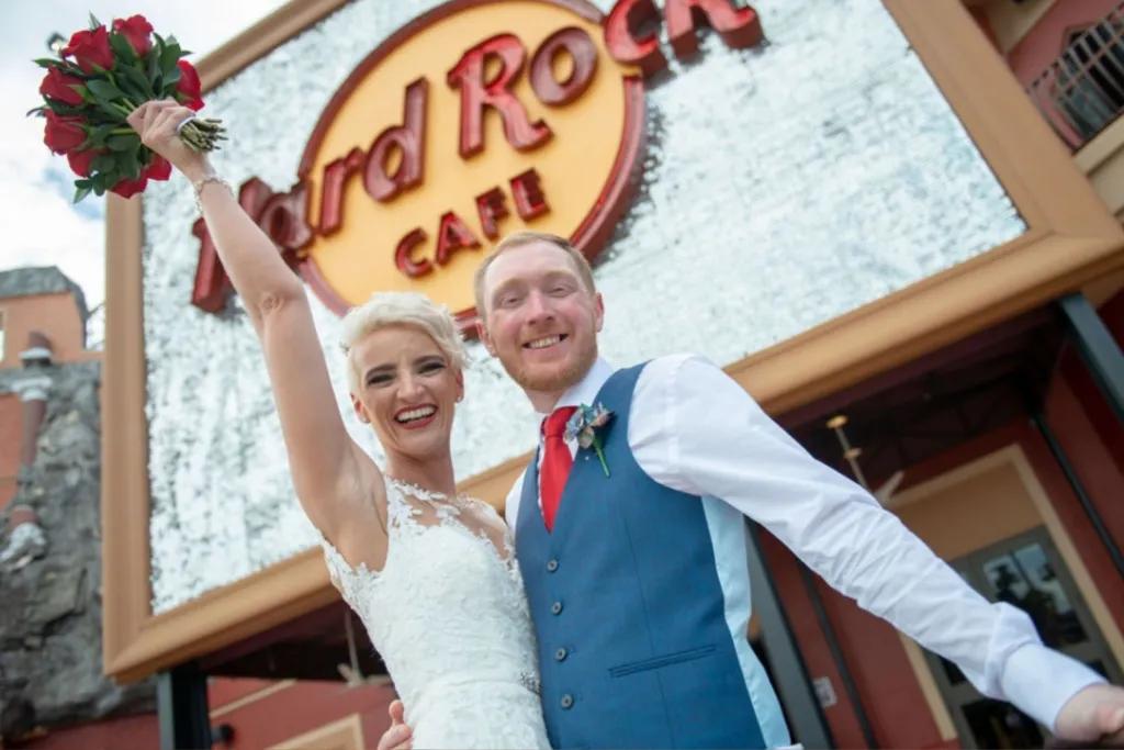 A bride holding her bouquet in the air with her arm around her groom. They are posing in front of the Hard Rock Cafe sign