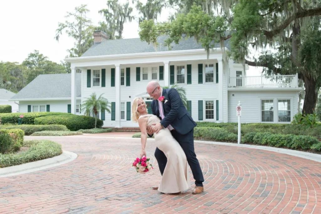 A groom dipping his bride over his knee. They are posing in front of a white American country house.
