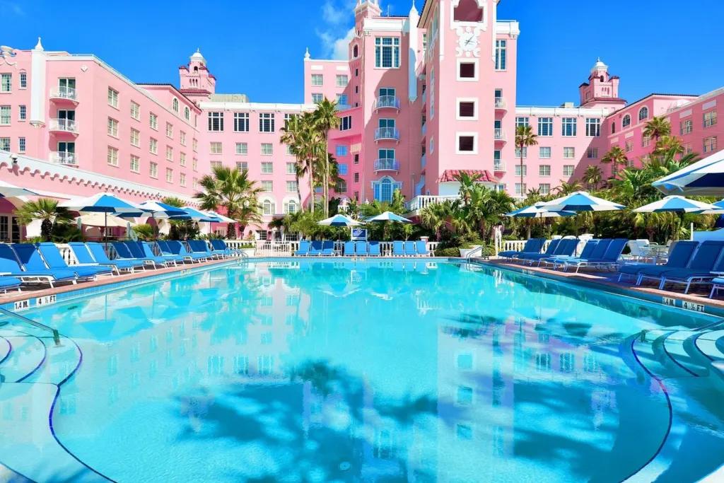 An empty hotel pool lined with blue sun loungers in front of a bright pink hotel building