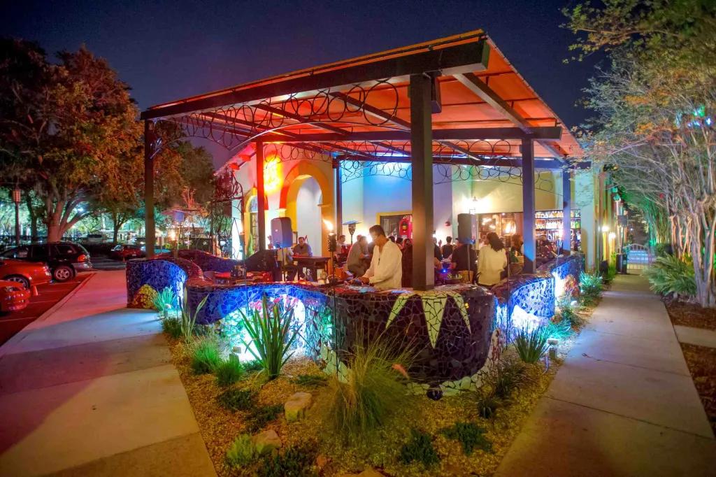 Large wooden beams holding up an orange roof and a mosaic blue wall winding around the terrace. There are speakers, heaters and people stood on the terrace and there is blue and orange lighting.