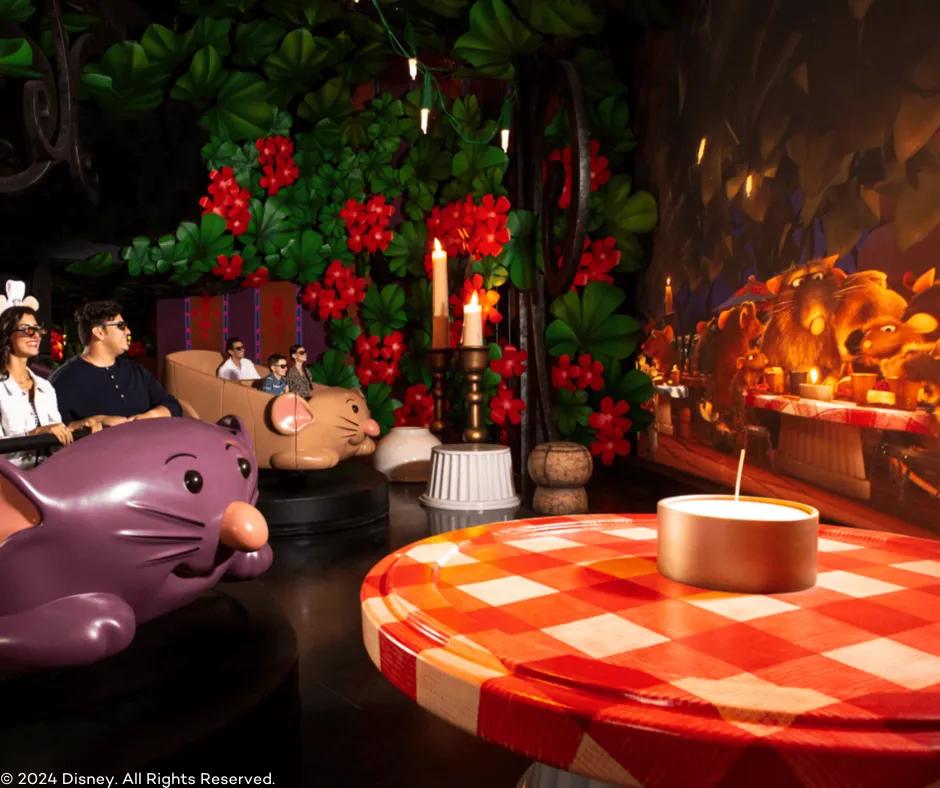 Guests sat in a rat shaped vehicle with 3D glasses on. Next to them on the right is a giant bush with large flowers, a giant table with a red and white table cloth, giant candles and a screen with the ratatouille characters on. 