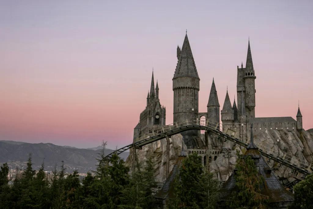 Grey Hogwarts castle sat on rocks with trees and small coaster in front. In the background there is a pink sunset and large mountains.