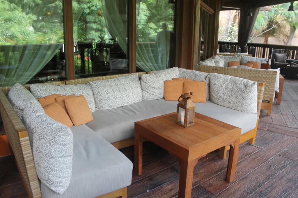 Terrace with a small, orange tinted wooden table with copper lantern on it surrounded by a white cushioned sofa with two corners. Sat in front of three large glass windows with wooden beams. In the background is more tables and trees.