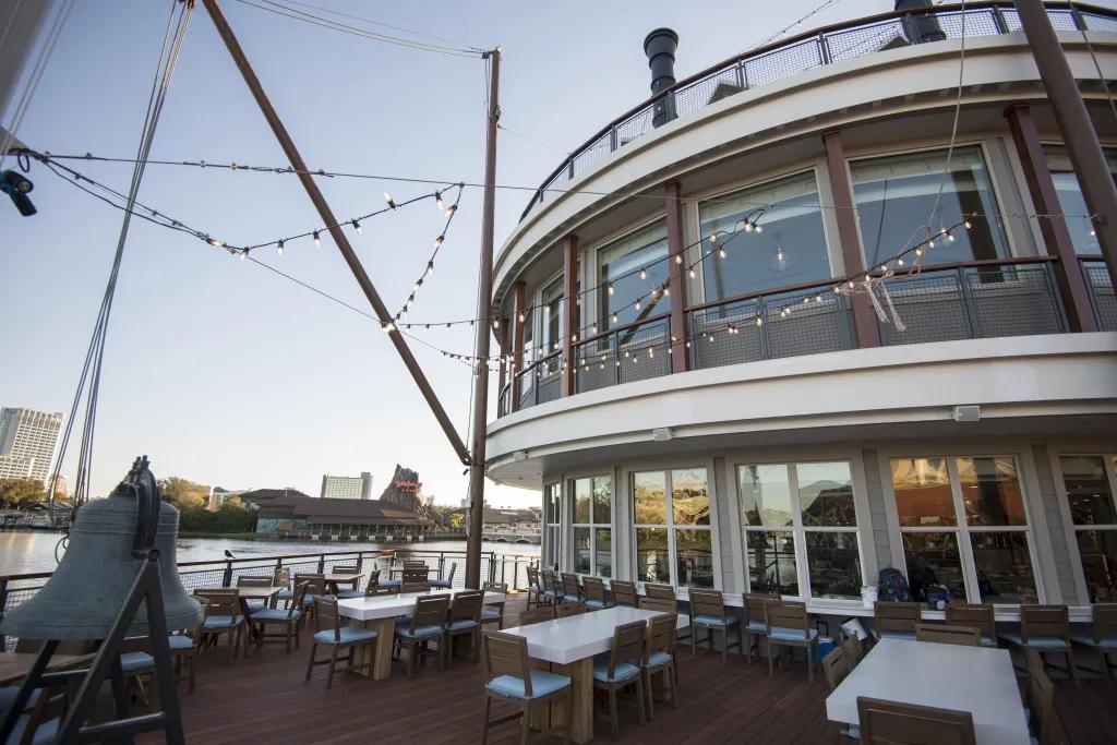 White tables with 6 wooden chairs around them on a terrace with lake in the background and lights hanging overhead. Also a large bell on the terrace and circular building to the right.