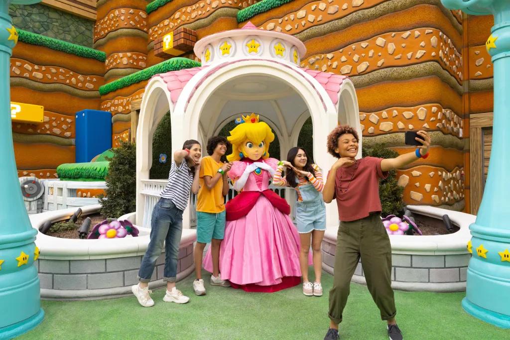 Princess Peach, a blonde character with pink princess dress, stood underneath a white and pink gazebo with four guests around her taking a picture with her. In the background is the brown land decoration.